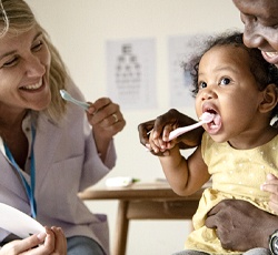 A dentist holding a toothbrush while a father holds his toddler and shows her how to brush her teeth